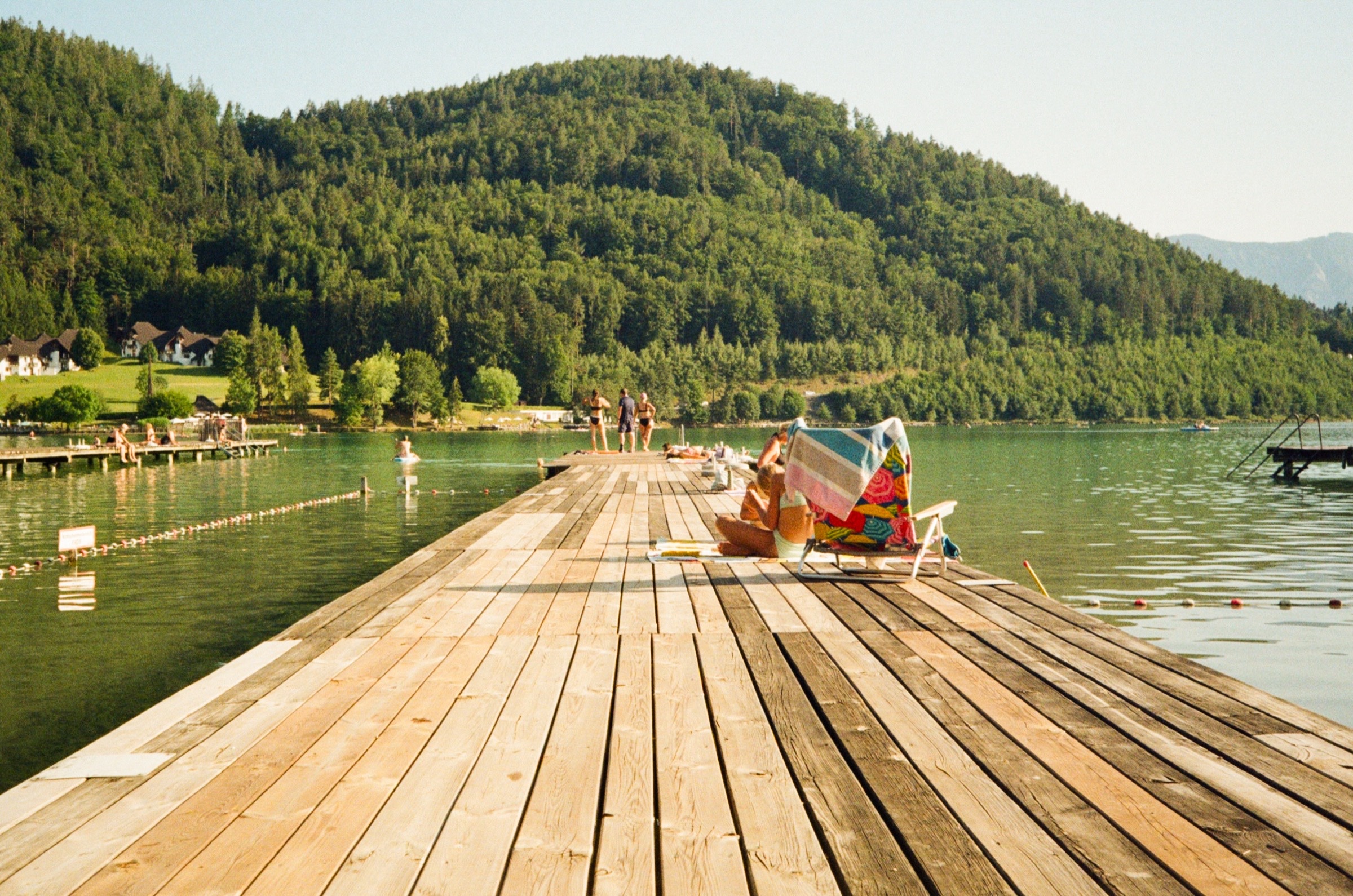 Wooden pier stretching into the turquoise water of Klopeiner See with forested mountains beyond, Austria - Travel photography by Rocco Goldschmidt