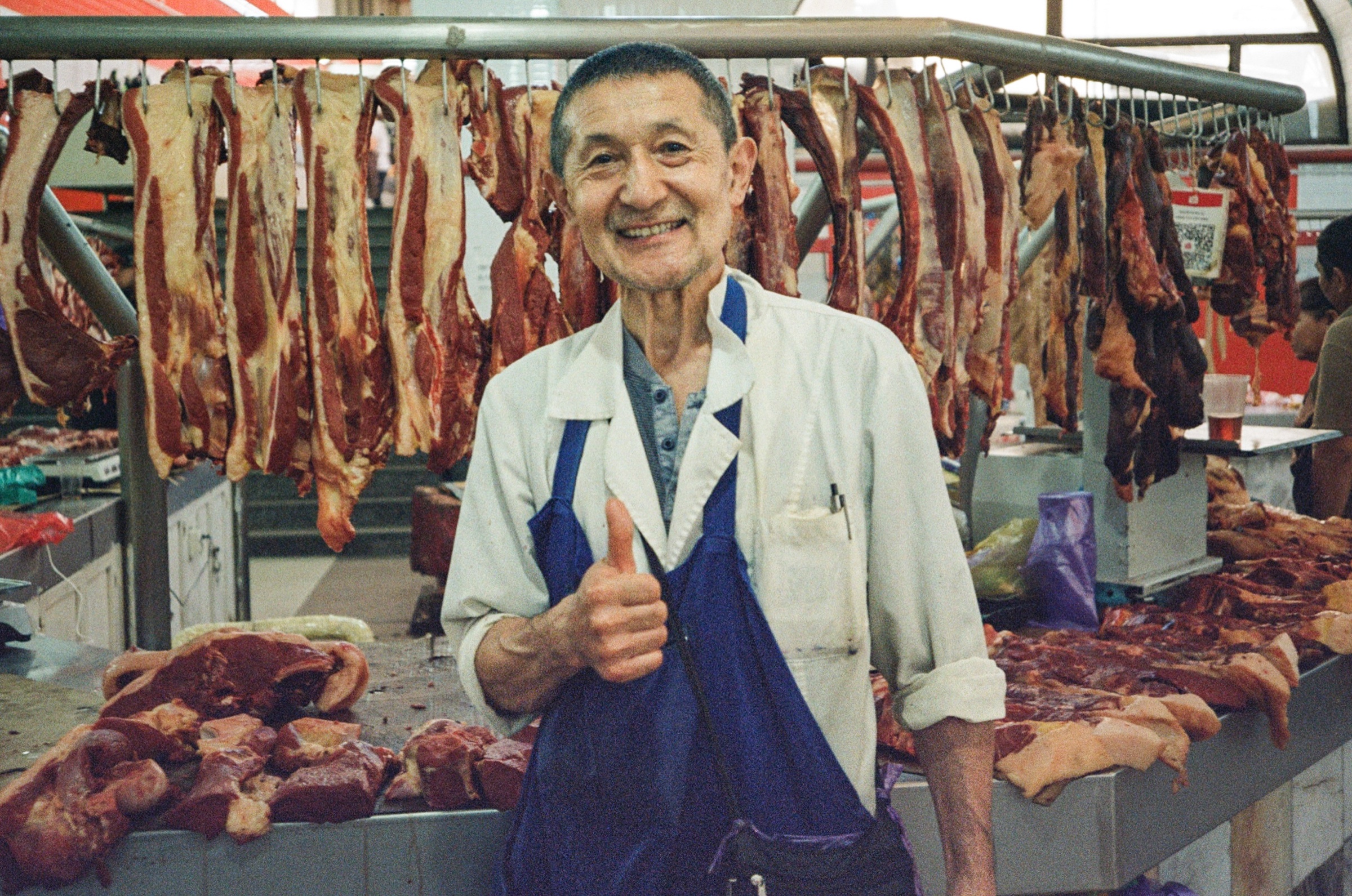 Smiling butcher giving a thumbs up in front of hanging cuts of meat at a market in Bishkek, Kyrgyzstan - Street portrait photography by Rocco Goldschmidt
