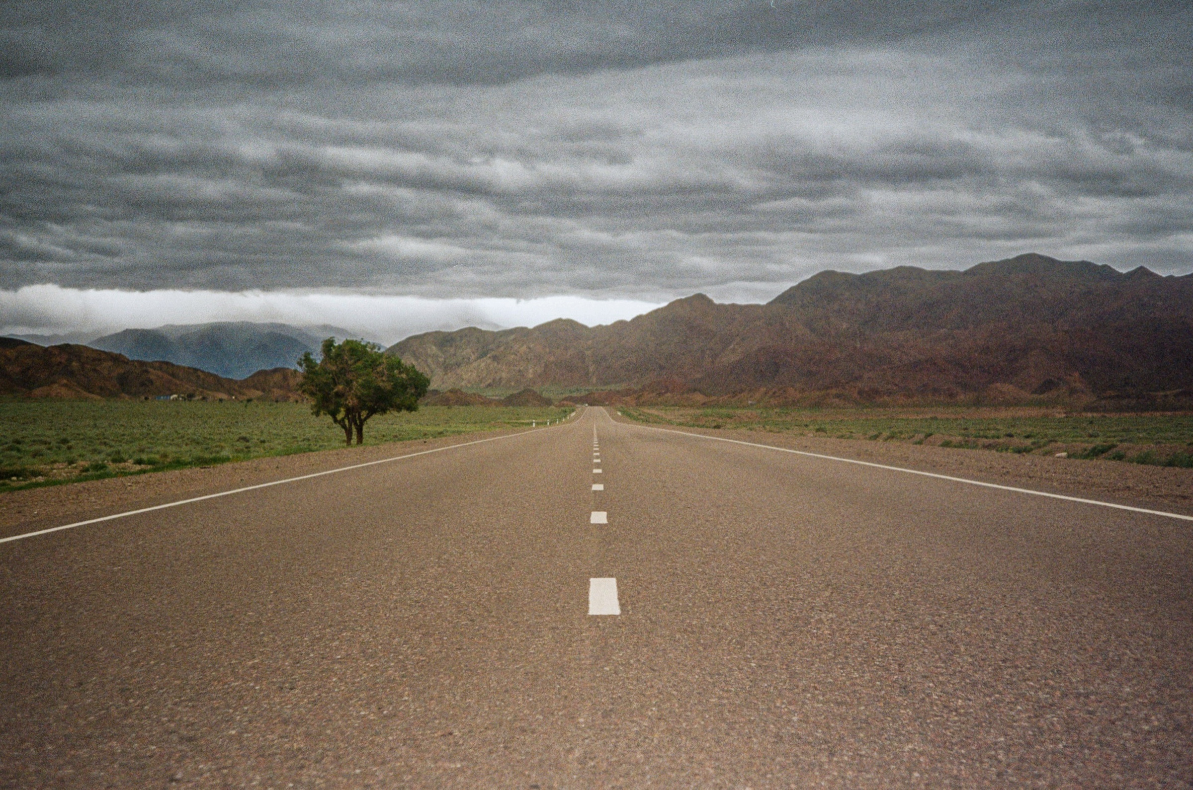 Empty highway stretching toward stormy mountains in the Naryn region, Kyrgyzstan - Travel photography by Rocco Goldschmidt
