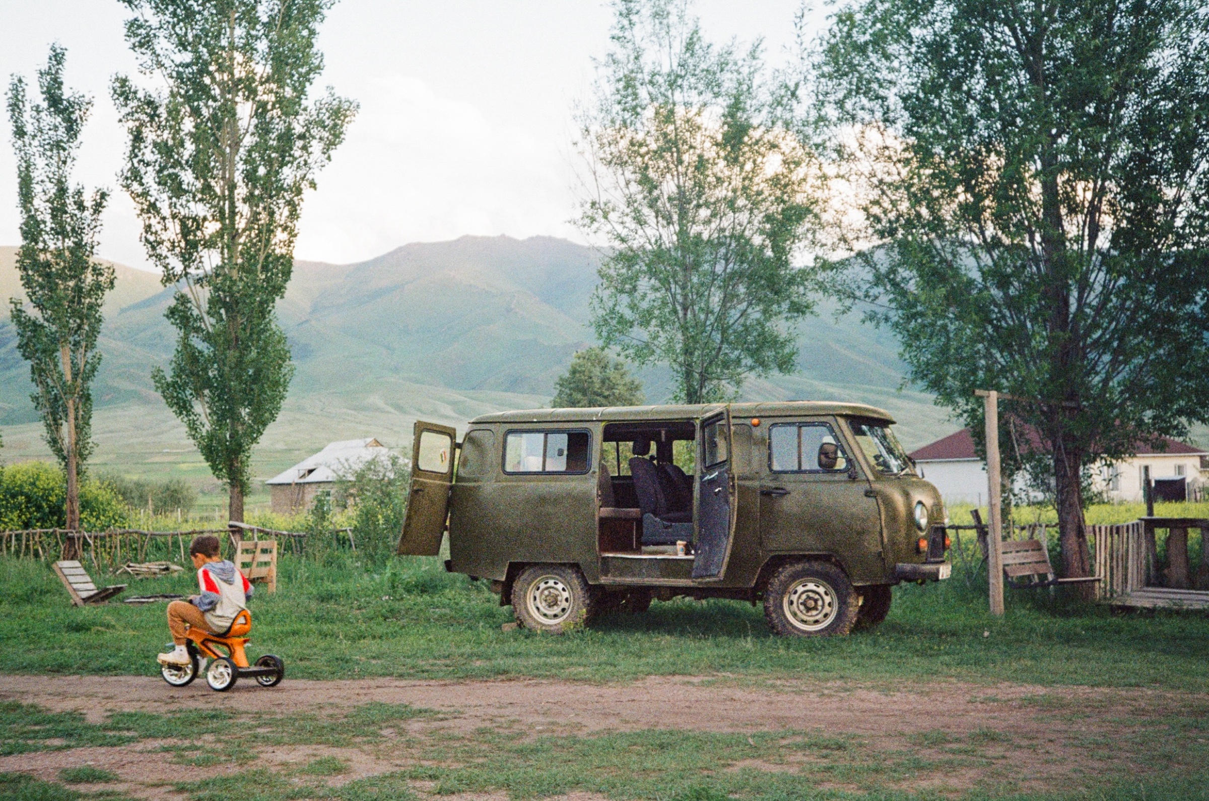 Child on a small tricycle beside a Soviet UAZ van at a mountain homestay in Kyzart, Kyrgyzstan - Street photography by Rocco Goldschmidt