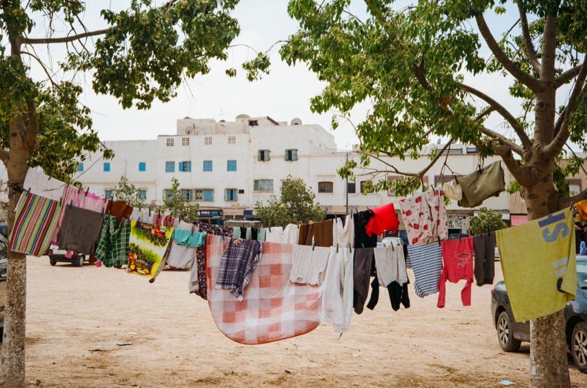 Man pushing a wheelbarrow across a sandy square flanked by white buildings in Agadir, Morocco - Street photography by Rocco Goldschmidt
