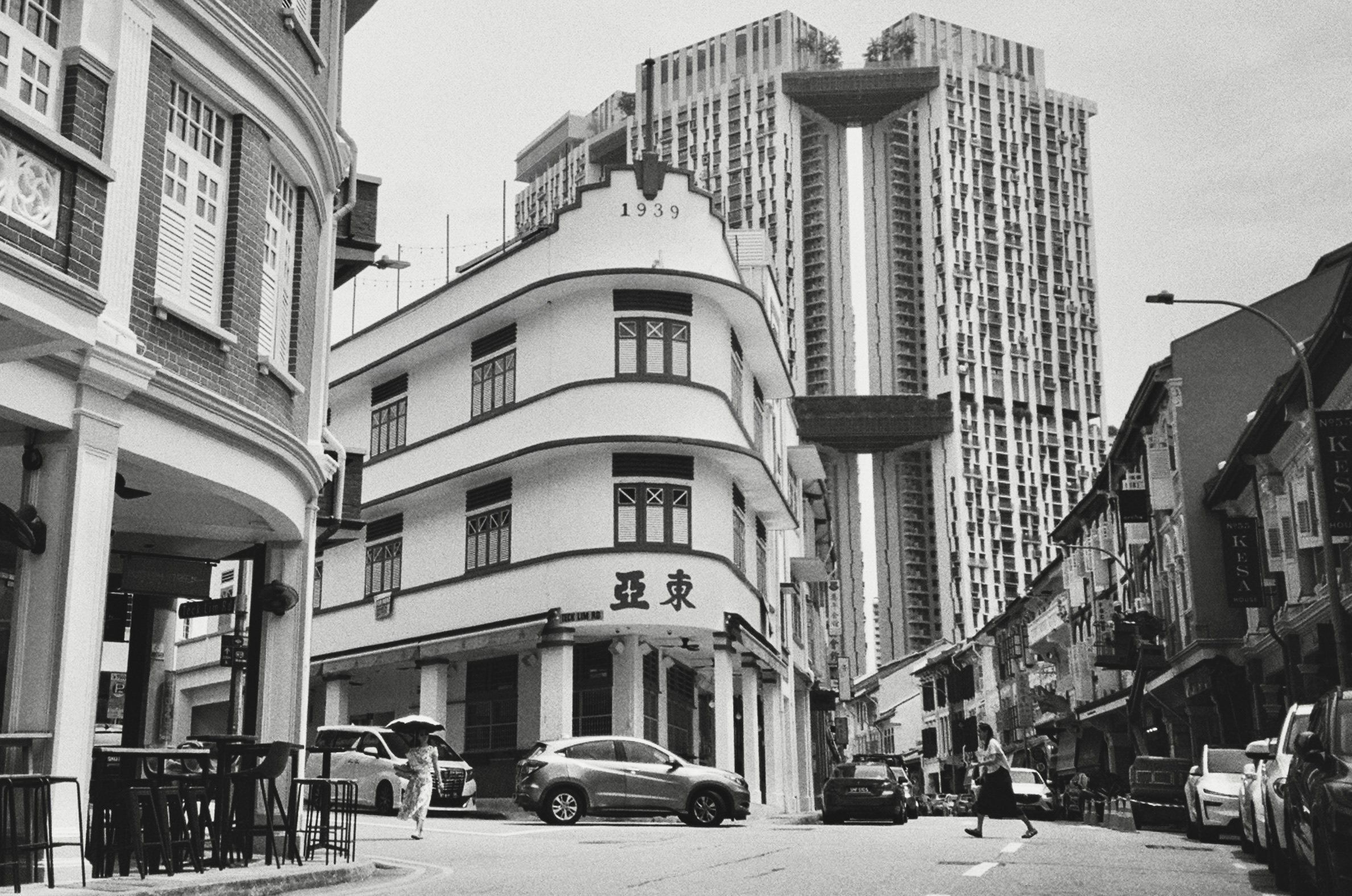 Black and white street corner with a curved 1939 shophouse in front of twin residential towers joined by sky bridges, a woman crossing with an umbrella - Film photography by Rocco Goldschmidt
