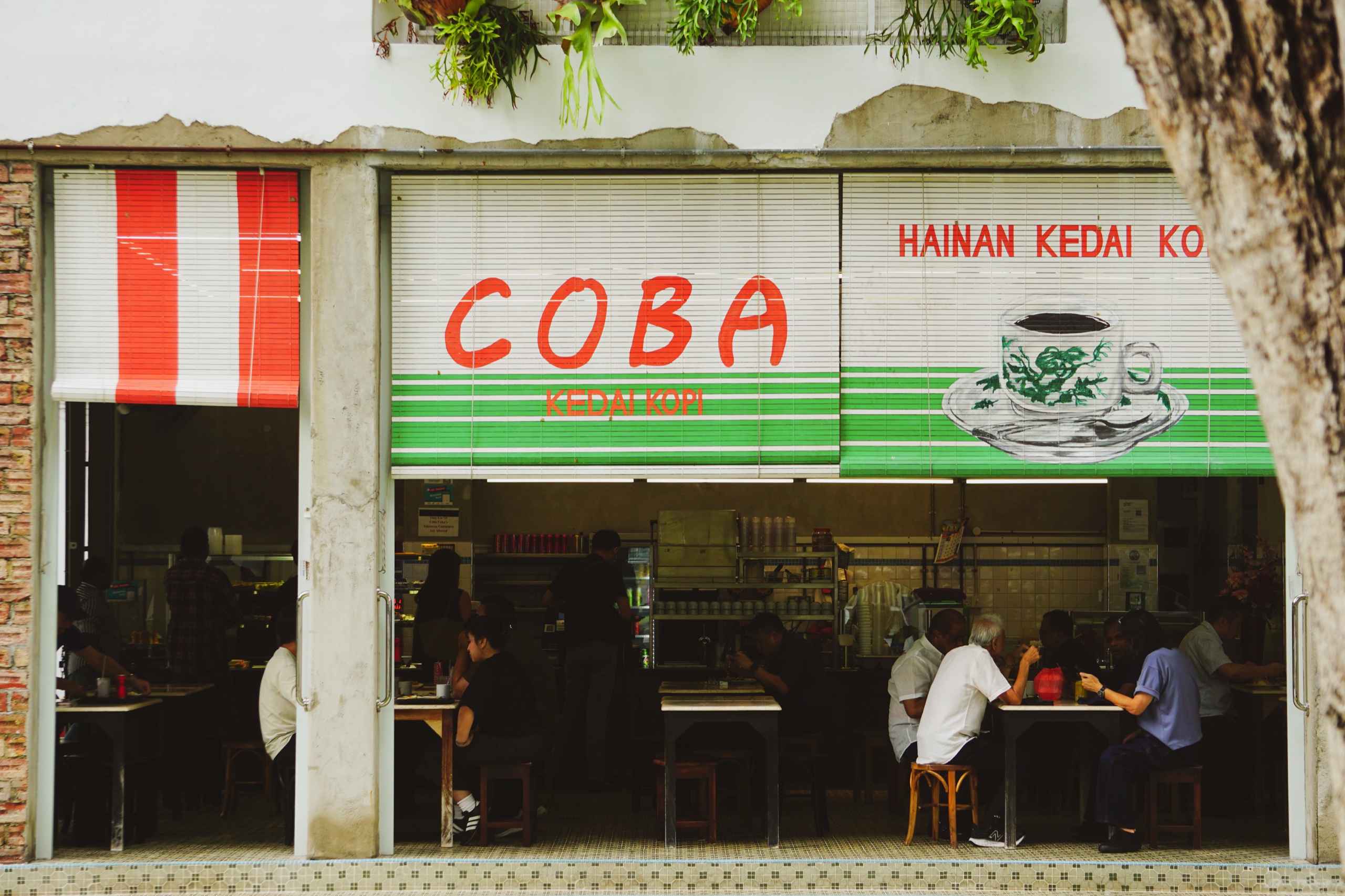 Coba Hainan kedai kopi with green and white painted blinds and a coffee cup sign, people eating at tables inside the open shopfront - Photography by Rocco Goldschmidt