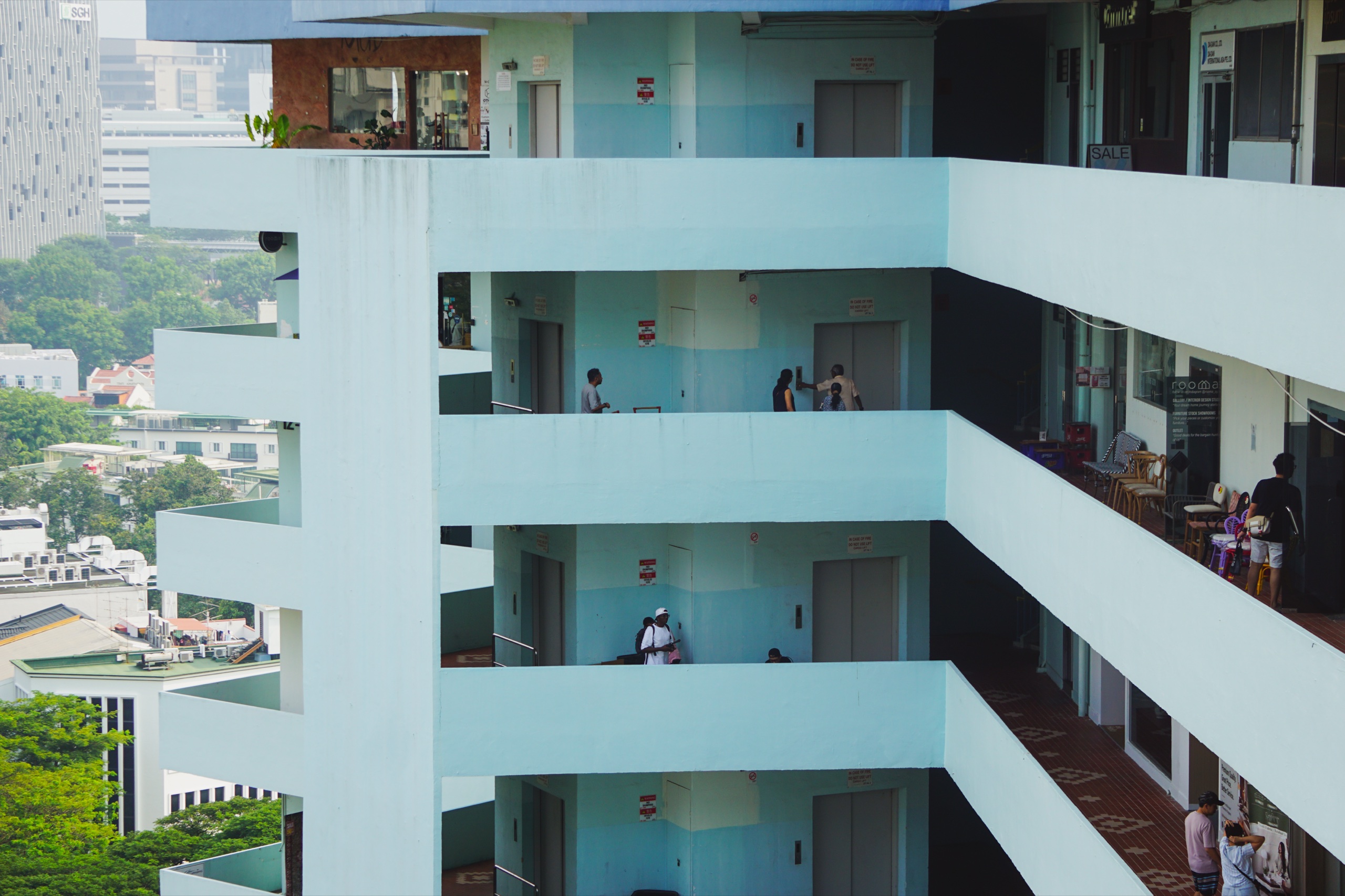 Pale mint-green open corridors stacked on several floors of a building, with people waiting by the lifts and greenery and towers in the distance - Photography by Rocco Goldschmidt