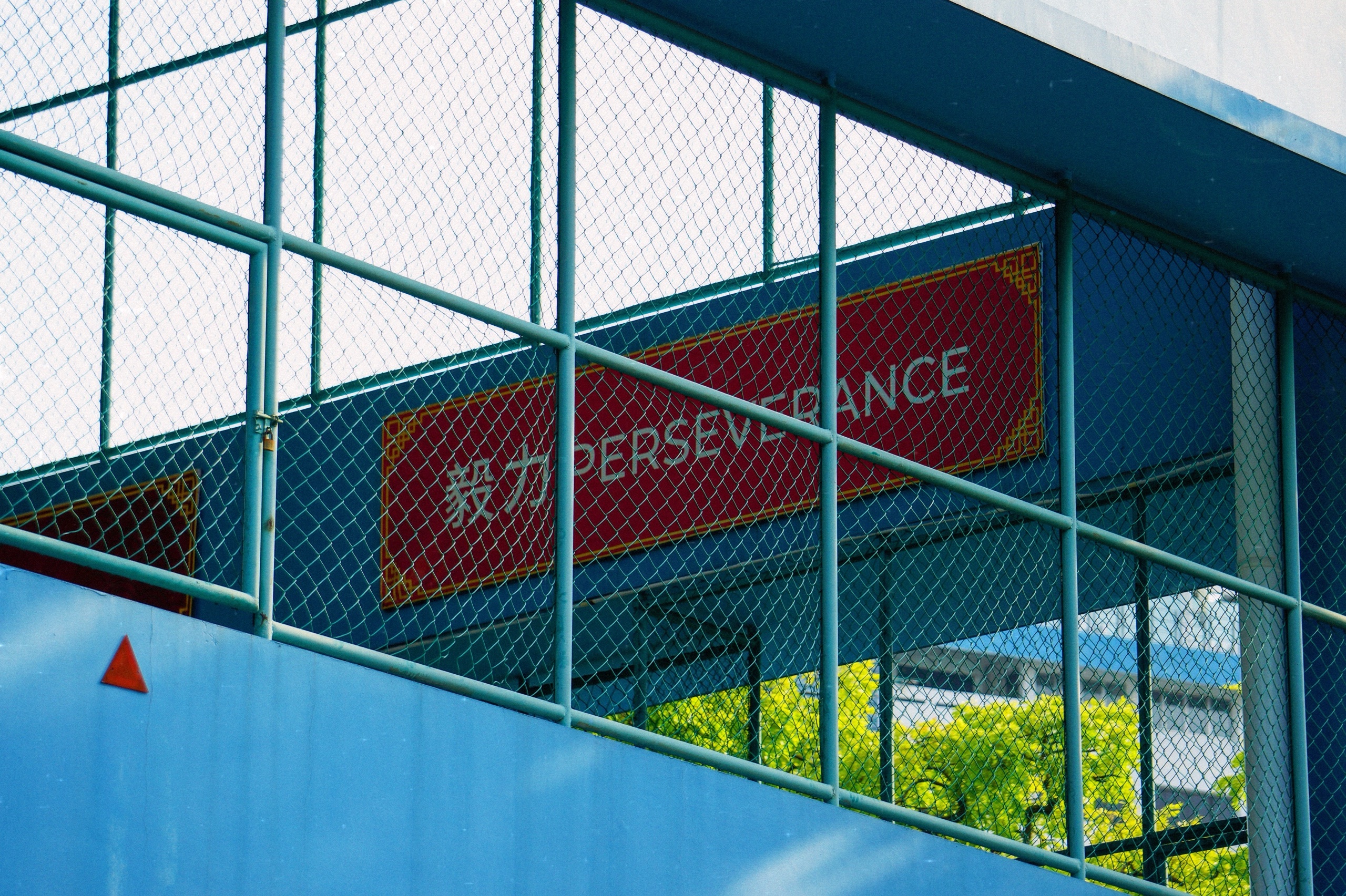 Red Perseverance sign in English and Chinese behind a chain-link fence on a blue building in Singapore - Film photography by Rocco Goldschmidt