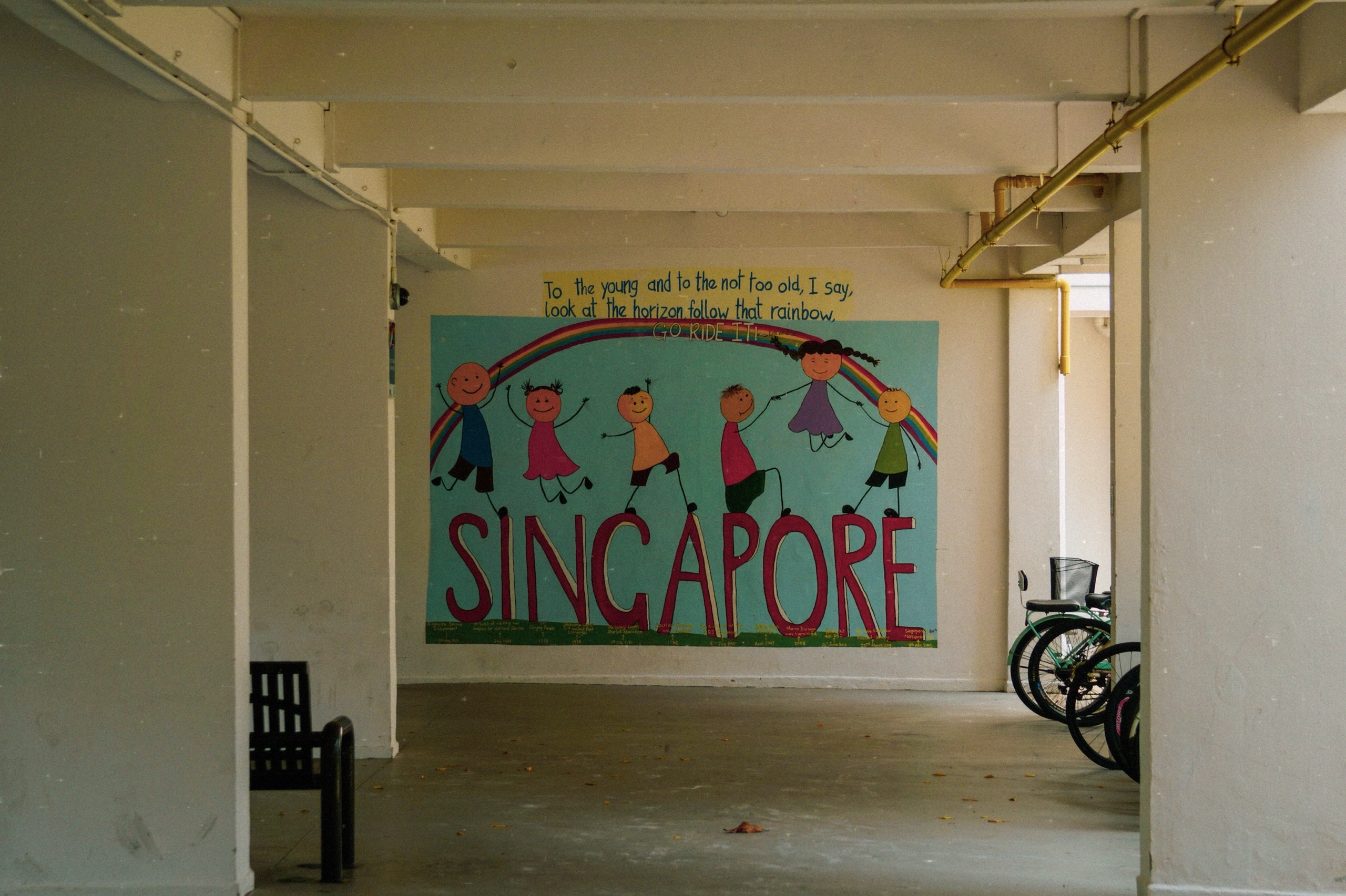 Children's rainbow mural spelling Singapore at the end of a quiet void deck, with bicycles parked beside the pillars - Film photography by Rocco Goldschmidt