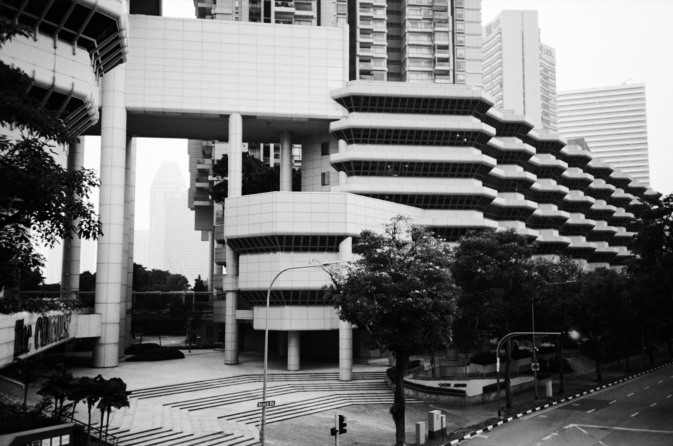 Black and white view of The Concourse, a stepped white tower on stilts with layered balconies above an empty plaza and tree-lined road - Film photography by Rocco Goldschmidt
