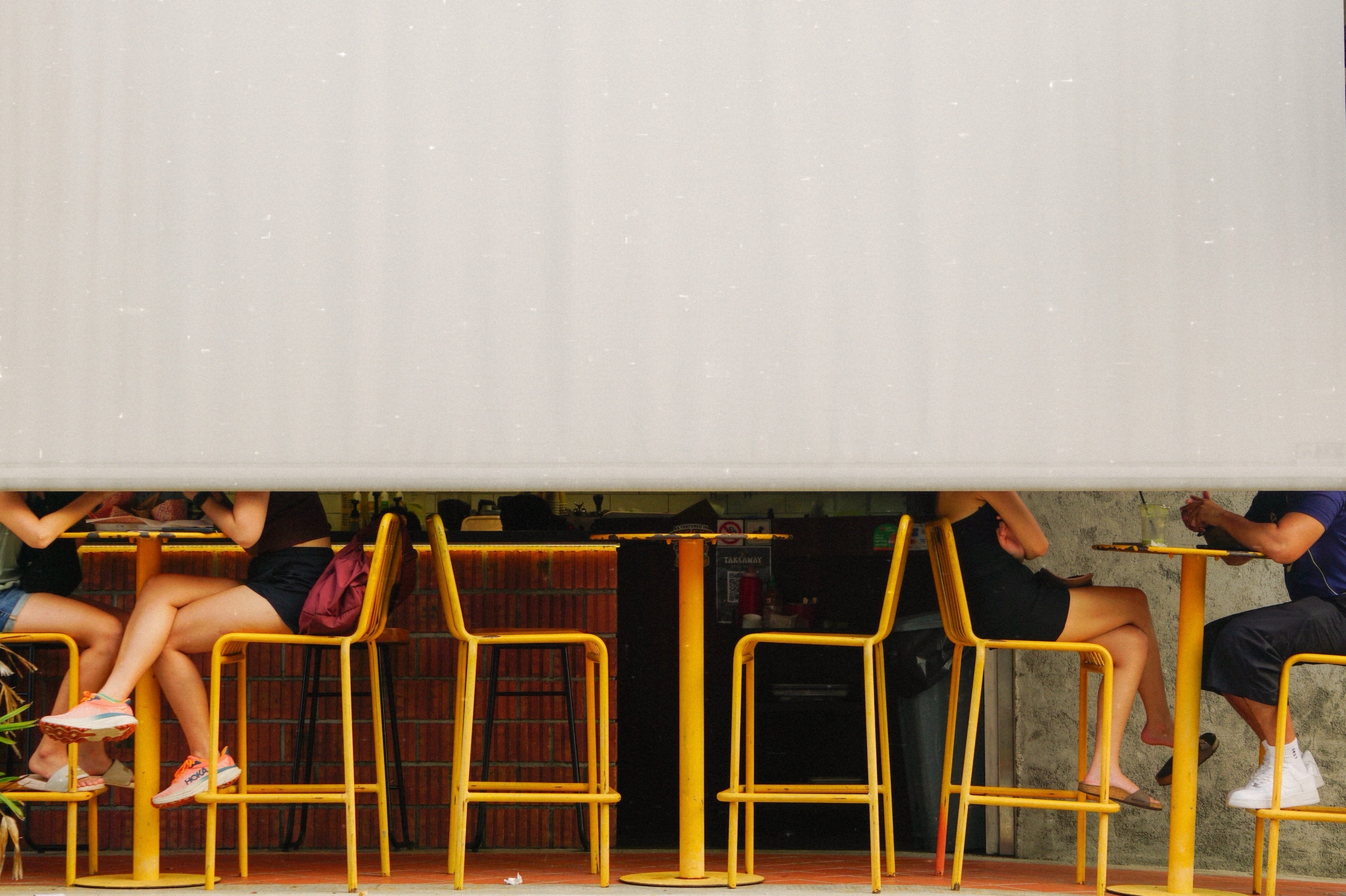 People seated on yellow bar stools below a half-lowered white shutter at a cafe counter in Singapore - Street photography by Rocco Goldschmidt