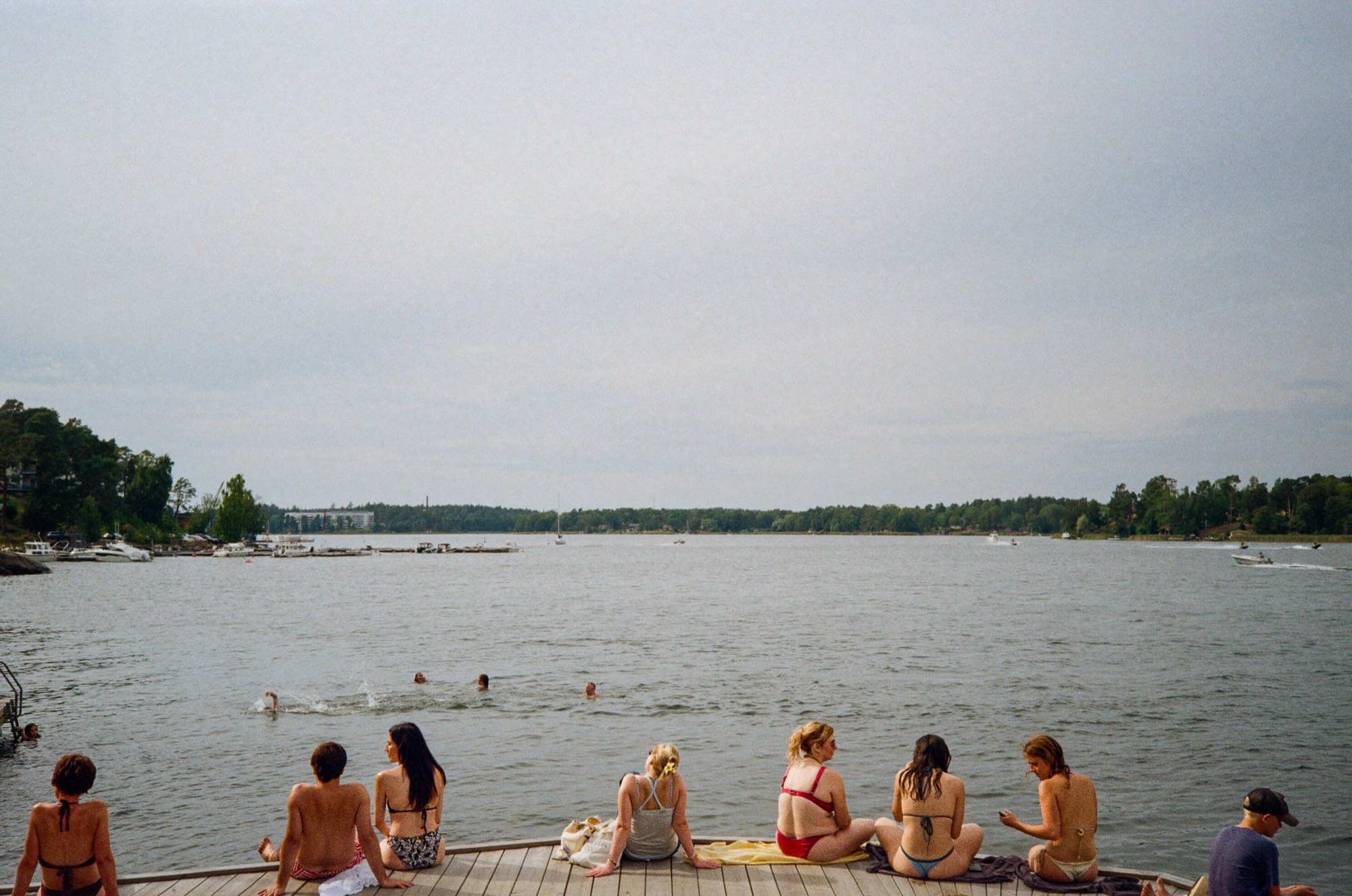 Friends sitting on a swimming dock watching bathers in the water on an overcast day in Stockholm, Sweden - Documentary photography by Rocco Goldschmidt